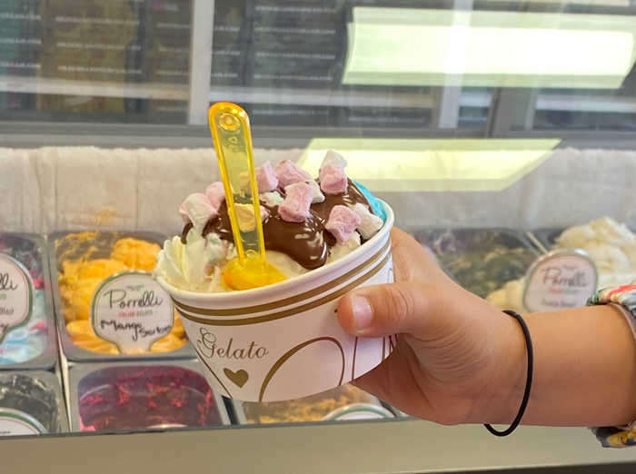 Hand holding a cup of Pirrelli Gelato topped with whipped cream, chocolate sauce, 
and mini marshmallows, purchased at Candy Cream Bathgate