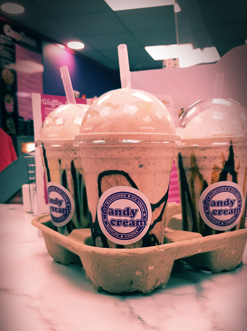 Three fresh milkshakes with chocolate drizzle and Candy Cream logo stickers, ready 
to be served in a carry tray at the Bathgate dessert shop