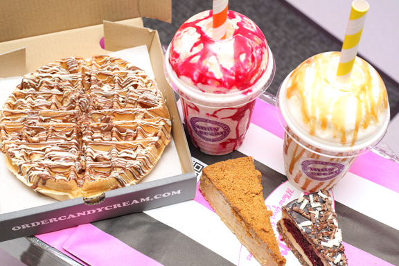 Gourmet milkshake drizzled with red syrup, standing behind slices of chocolate, 
Biscoff, and fruit cheesecake from Candy Cream Bathgate